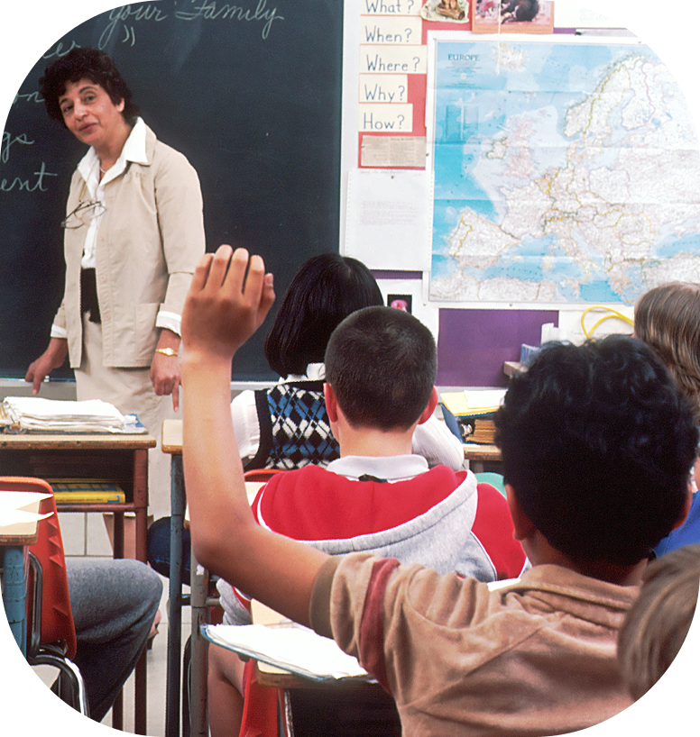 Image of a teacher giving a lesson in front of a blackboard. A student is raising his hand.